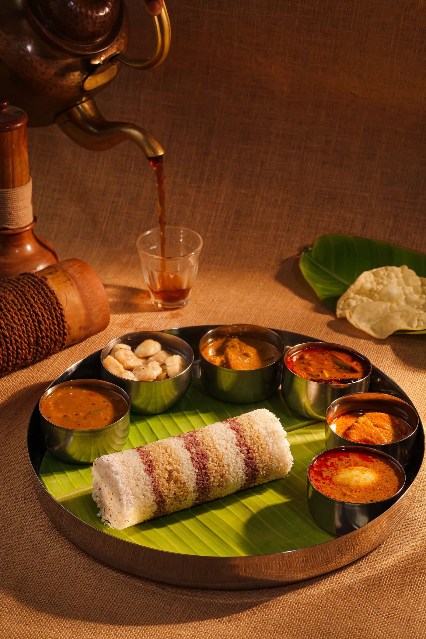 A puttu thali on a steel platter, puttu on a banana leaf ringed by small bowls of curry, while tea is poured behind